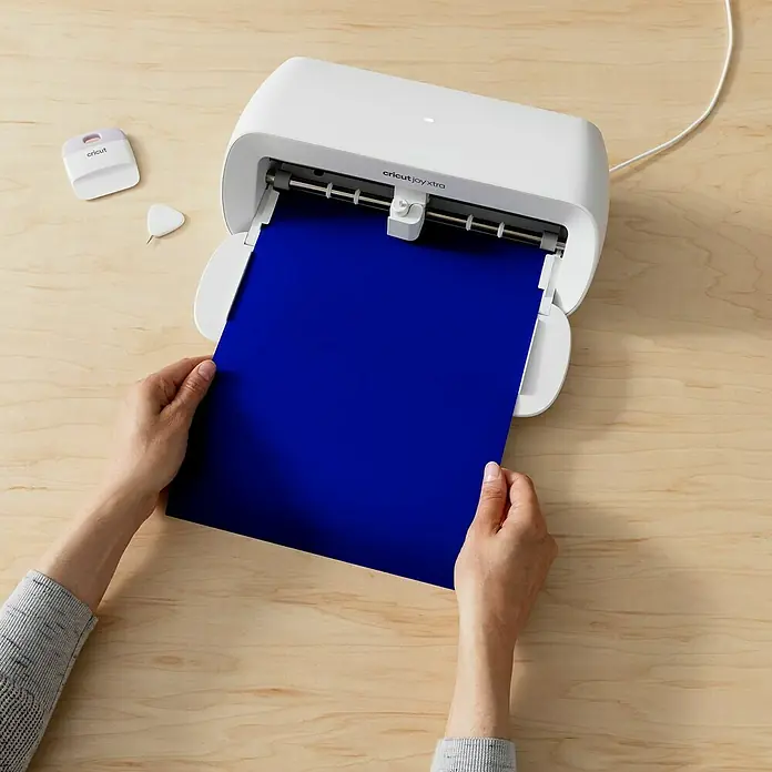 Cricut Joy Xtra Vinylfolie Smart Permanent - 3 -tlg., Pinwheel, 24,1 x 30,5 cm - Ein Cricut Joy Xtra Schneideplotter wird mit einem blauen Papierstück gefüttert, Werkzeuge liegen daneben.