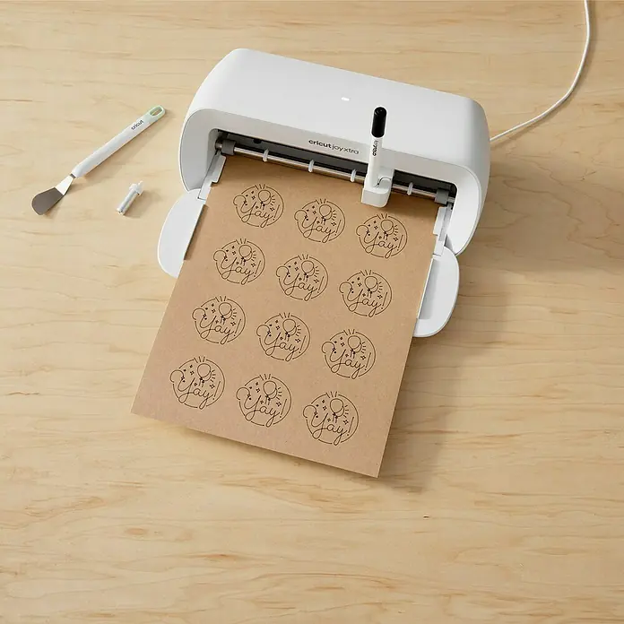 Cricut Joy Xtra Aufkleber Smart Label Paper Permanent - 4 Stk., Braun, 24,1 x 30,5 cm - Nahaufnahme eines Cricut Joy Xtra Bastelplotters beim Zeichnen von "Yay!"-Designs auf braunes Papier, Werkzeuge liegen daneben auf einem Holztisch.
