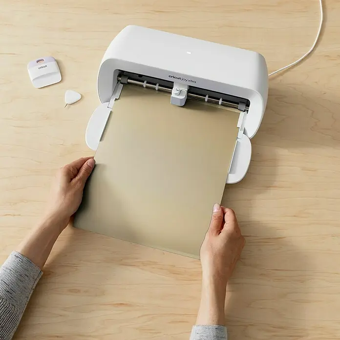 Cricut Joy Xtra Vinylfolie Smart Ablösbar - 3 -tlg., Classics, 24,1 x 30,5 cm - Nahaufnahme eines Cricut Joy Xtra DIY-Plotters mit einer Person, die eine Schneidematte einlegt, Werkzeuge liegen daneben auf einem Holztisch.