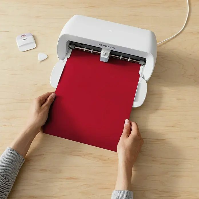 Cricut Joy Xtra Aufbügelfolie Smart Iron-On - Rot, 1 Stk., L x B: 61 x 24,1 cm - Nahaufnahme eines Cricut Joy Xtra-Schneideplotters mit rotem Papier, umgeben von Werkzeugen auf einem Holztisch.