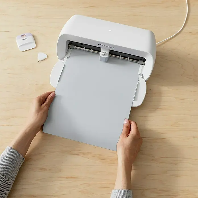 Cricut Joy Xtra Vinylfolie Smart Metallisch Permanent - 1 Stk., Silber, 24,1 x 91,4 cm - Eine Cricut Joy Xtra Schneidemaschine mit hellgrauem Papier wird auf einem hellen Holztisch verwendet, Werkzeuge liegen daneben.