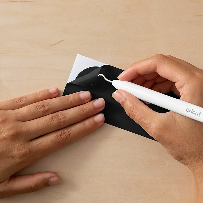 Cricut Joy Xtra Vinylfolie Smart Permanent - 3 -tlg., Elegance, 24,1 x 30,5 cm - Eine Person, die schwarzes Vinyl mit einem weißen Cricut Werkzeug bearbeitet, um unerwünschte Teile zu entfernen.
