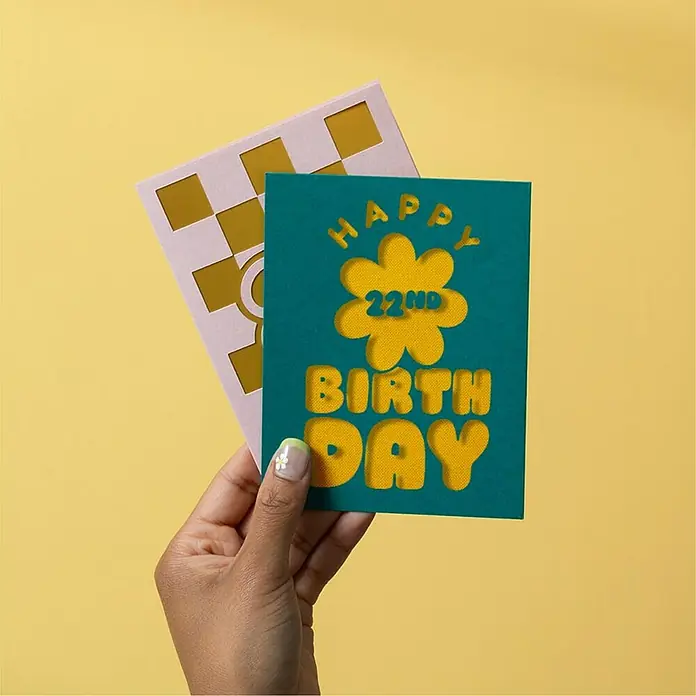 Cricut Joy Xtra Schneideplotter Starter Bundle - Weiß, Bluetooth, Schnittbreite: 21,6 cm, Schnittlänge: 1,2 m - Eine Hand hält eine Geburtstagskarte zum 22. Geburtstag mit gelben Ausschnitten vor einem gelben Hintergrund.