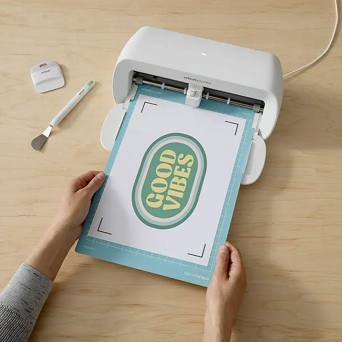 Cricut Aufbügelfolie Light Fabrics - 5 Stk., Weiß, L x B: 29,7 x 21 cm - Ein Cricut Joy Xtra Schneideplotter in Weiß schneidet ein Blatt mit dem Aufdruck "Good Vibes" auf einer Schneidematte.