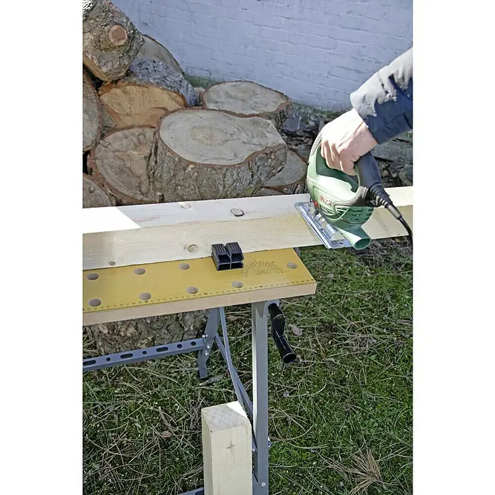 Werkbank Klappbar - L x B: 63,5 x 60,6 cm, Arbeitshöhe: 76 cm, Belastbarkeit: 150 kg - Eine grüne Stichsäge schneidet ein Stück Holz auf einer gelben Werkbank mit einem Stapel Baumstämme im Hintergrund.