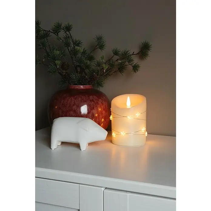 Konstsmide LED-Kerze - Cremeweiß, Ø x H: 10 x 14 cm, Echtwachs - Dekorative LED-Kerze mit Sternenlicht und eine weiße Figur auf einer Kommode, neben einer Vase mit Tannenzweigen für gemütliche Atmosphäre.