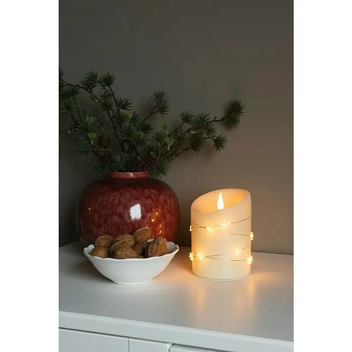 Konstsmide LED-Kerze - Cremeweiß, Ø x H: 10 x 14 cm, Echtwachs - Kerze mit Sternenlichtern neben einer Vase mit Tannenzweigen und einer Schale mit Walnüssen auf einem weißen Tisch.
