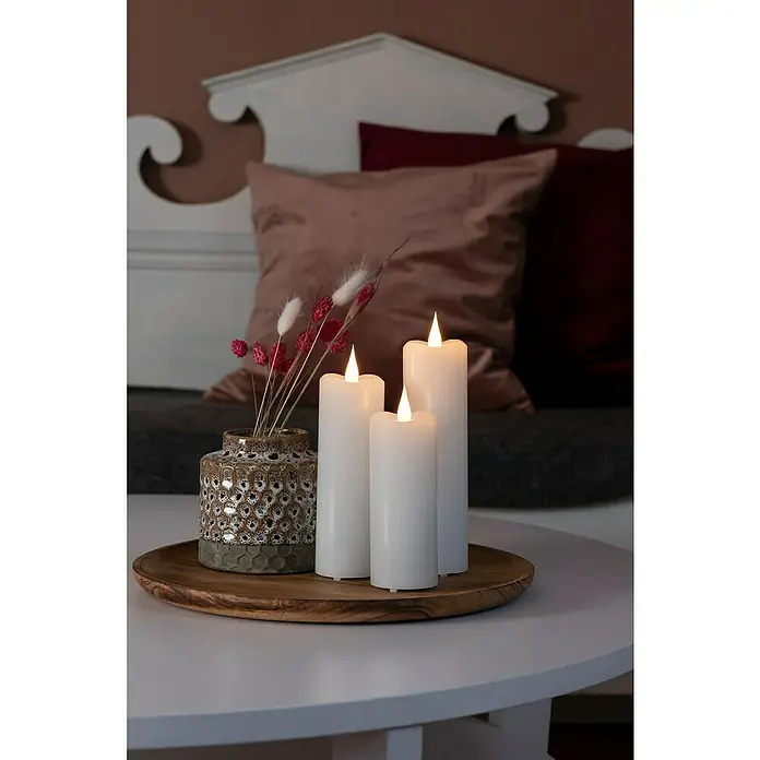 Konstsmide LED-Kerze - Weiß, Ø x H: 7,8 x 22 cm, Echtwachs - Nahaufnahme von dekorativen LED-Kerzen und einer Vase auf einem Holzteller, die eine warme, gemütliche Atmosphäre schaffen.