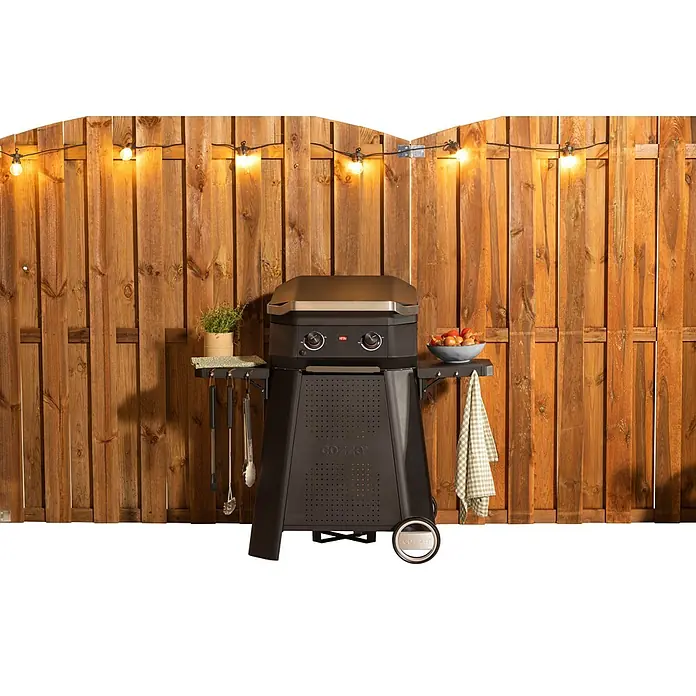 Cozze Elektrogrill E 500 - Grillfläche (B x T): 51 x 39 cm, 2,1 kW, Schwarz - Ein Gozney Dome Ofen mit Reglern, Ablageflächen und Zubehör vor einem Holzzaun mit Lichterkette.