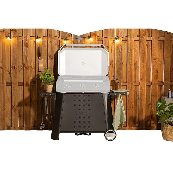 Cozze Rollwagen - B x T x H: 146 x 70 x 74,5 cm, Stahl, Schwarz, Passend für: Cozze Plancha Grill 800 - Bild eines Cozze Grills mit geöffnetem Deckel vor einem Holzzaun, dekoriert mit Lichterketten und Zubehör.