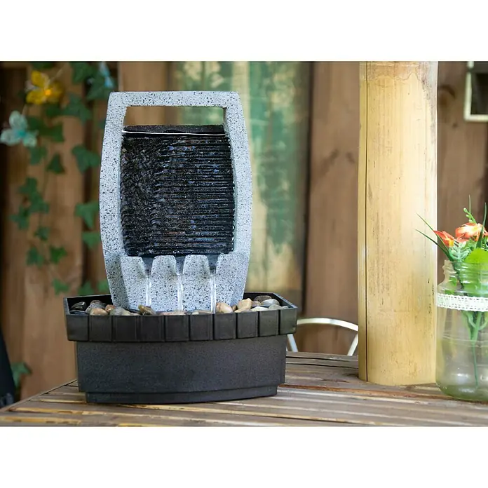 Dobar Gartenbrunnen - 20,5 x 16,5 x 28 cm, Kunststoff, Grau/Schwarz - Nahaufnahme eines Zimmerbrunnens mit Wasserfall-Design, der auf einem Holztisch steht und von einer Vase mit Blumen begleitet wird.