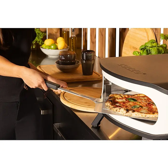 Cozze Pizzaofen mit drehbarem Pizzastein - Durchmesser Grillfläche: 42,5 cm, 6 kW, Weiß/Schwarz, Stahl - Eine Person schiebt eine Pizza mit einem Pizzaschieber in einen Cozze Pizzaofen, der auf einer Küchentheke steht.