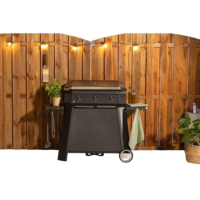 Cozze Plancha-Grill 800 - Grillfläche (B x T): 80 x 50 cm, 7,5 kW, Schwarz - Cozze Gasgrill mit Zubehör vor Holzzaun mit Lichterkette. Der Grill ist schwarz mit silbernen Akzenten und hat drei Brenner.