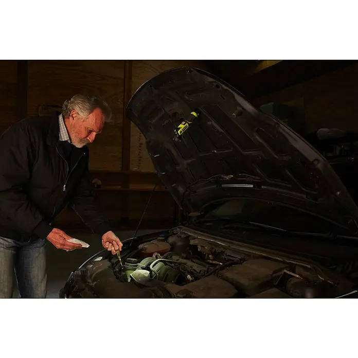 Ryobi 4V USB Akku-Taschenlampe RLP4-0 - 4 V, Ohne Akku - Ein Mann prüft den Ölstand seines Autos mit einem gelben Peilstab und einem Tuch in einer dunklen Garage, beleuchtet von einer Lampe unter der Motorhaube.