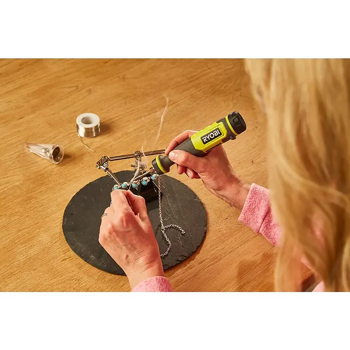 Ryobi 4V USB Akku-Lötstift RSI4-120G - 4 V, 1 Akku, 2 Ah - Eine Person, die mit einem Lötkolben der Marke Ryobi an einem Schmuckstück arbeitet, wobei Rauch aufsteigt.