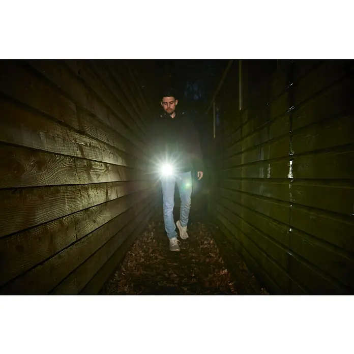 Ryobi 4V USB Akku-Taschenlampe RLP4 - 4 V, 1 Akku, Lichtstrom: 640 lm - Ein Mann in dunkler Kleidung leuchtet mit einer hellen Taschenlampe einen dunklen Gang aus Holz aus. Die Lampe wirft einen starken Schein.