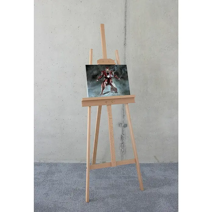 Komar Leinwandbild - Iron Man Charging, B x H: 40 x 30 cm - Nahaufnahme eines Holzstaffeleis mit einem Bild von Iron Man darauf, vor einer grauen Wand.