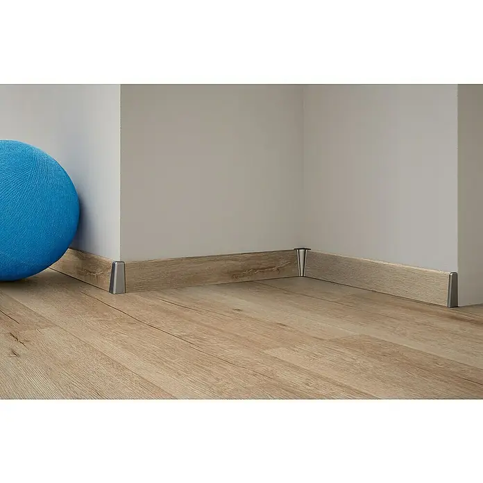 Laminado Roble Serena - 1.380 x 193 x 12 mm, AC5 - Toma detallada de un zócalo de madera con adornos esquineros plateados contra una pared clara y un piso de madera clara con una bola azul.