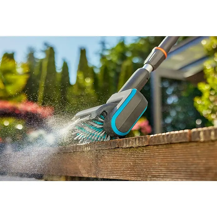 Gardena Power for All 18V Akku-Reinigungsbürste AquaBrush Patio 03/18V - 1 Akku - Eine Gardena Bürste säubert mit Wasser einen Holzbalken, während Spritzer in der Luft zu sehen sind.
