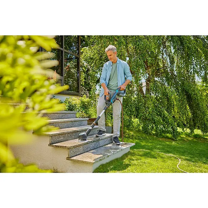 Gardena Power for All 18V Akku-Reinigungsbürste AquaBrush Compact 02/18V - Ohne Akku - Ein Mann reinigt mit der GARDENA Bürste PowerBrush die Außentreppe, umgeben von Grün. Das Gerät sorgt für saubere Steine im Garten.