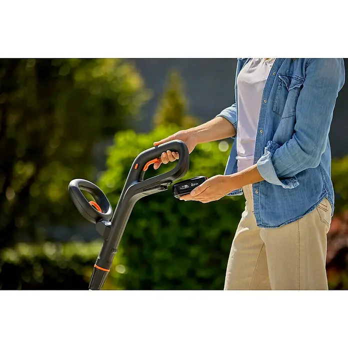 Gardena Power for All 18V Limpiador de juntas de batería EasyWeed 1800/18V - 18 V, 1 batería - Mujer reemplazando la batería de un recortabordes Gardena. El dispositivo es negro y naranja. La batería es pequeña y compacta con 2.0 Ah.