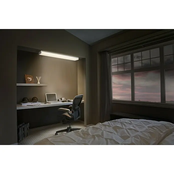 Ledvance LED-Deckenleuchte Office Line Wrap - 40 W, L x B x H: 120 x 16,31 x 7,16 cm, Weiß, Kaltweiß - Bürostuhl vor einem Schreibtisch mit Laptop und Accessoires, neben einem Bett und Fenster mit Aussicht.