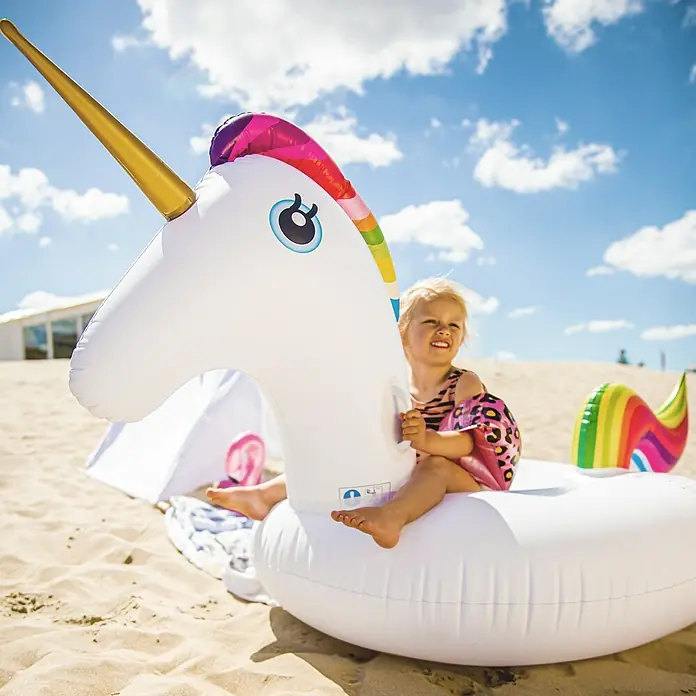 Swim Essentials Schwimmtier Einhorn - 142 x 137 x 97 cm - Ein fröhliches Kind sitzt auf einem aufblasbaren Einhorn am Strand, umgeben von Sand und blauem Himmel mit Wolken.