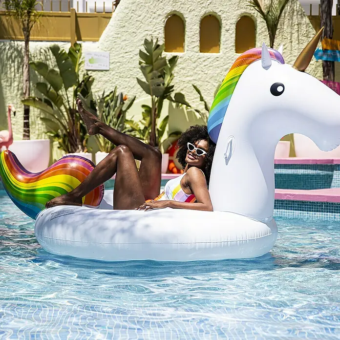 Swim Essentials Schwimmtier Einhorn - 142 x 137 x 97 cm - Eine Frau entspannt sich auf einer aufblasbaren Einhorn-Luftmatratze im Pool, die mit einem bunten Regenbogen-Design versehen ist.