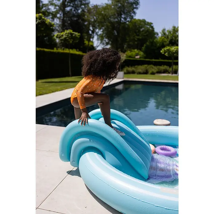 Swim Essentials Planschbecken - 203 x 173 x 89 cm, Regenbogen - Ein kleines Mädchen in einem orangefarbenen Badeanzug klettert auf eine blaue aufblasbare Wasserrutsche am Pool. Ideal für sommerlichen Wasserspaß.