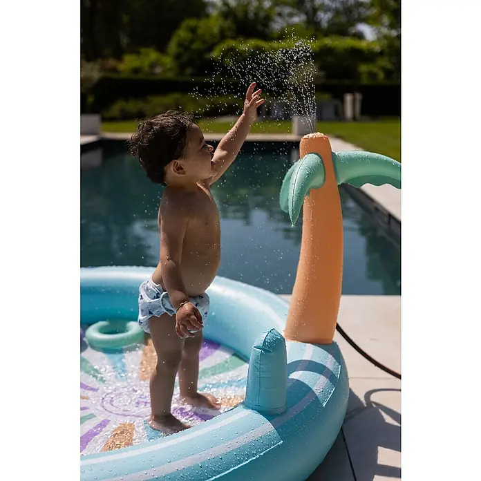 Swim Essentials Planschbecken - 203 x 173 x 89 cm, Regenbogen - Ein Kleinkind spielt im Planschbecken mit einer aufblasbaren Palme, die Wasser versprüht, an einem sonnigen Tag.