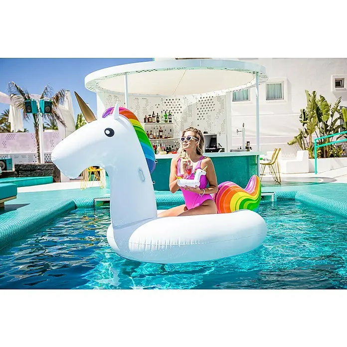 Swim Essentials Schwimmtier Einhorn - 142 x 137 x 97 cm - Frau genießt den Sommer auf einer aufblasbaren Einhorn-Luftmatratze im Pool, während sie einen Cocktail trinkt.