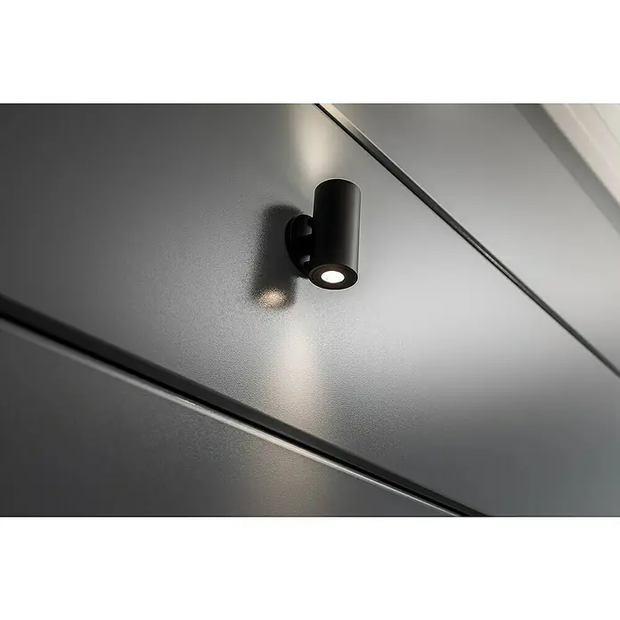 Telluria Pavillon Singolo 3630 - Außenmaß inkl. Dachüberstand (B x T): 388 x 328 cm, Stahl, Anthrazit, Ohne Set - Nahaufnahme einer schwarzen, zylindrischen Wandleuchte, die ein warmes Licht auf eine graue Oberfläche wirft.