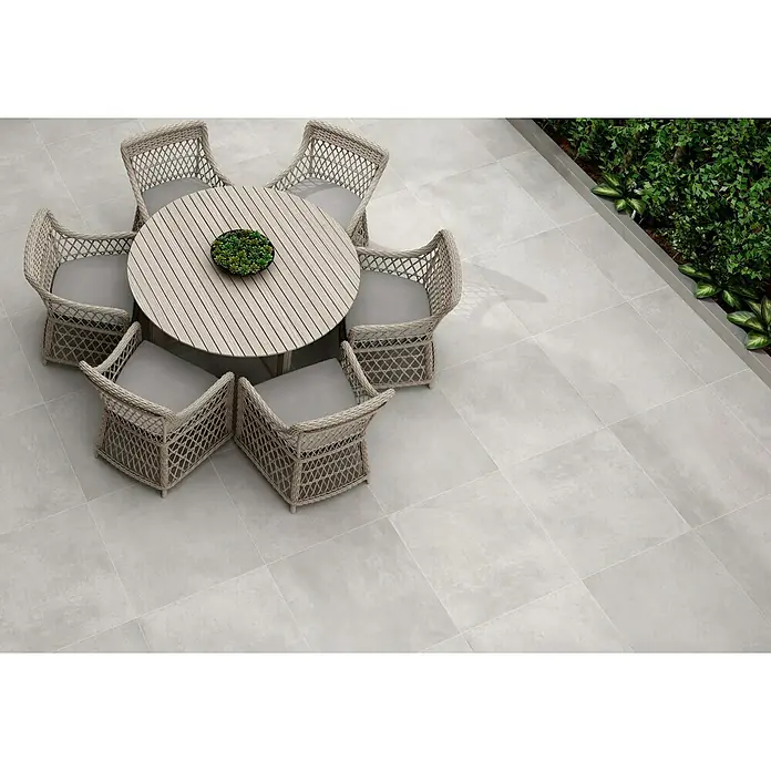 Pavimento porcelánico Rodano - 60 x 60 cm, Light grey, Mate - Atractiva zona de estar en el jardín con una mesa redonda de madera y seis sillas de mimbre sobre baldosas de color gris claro, ideal para relajarse al aire libre.