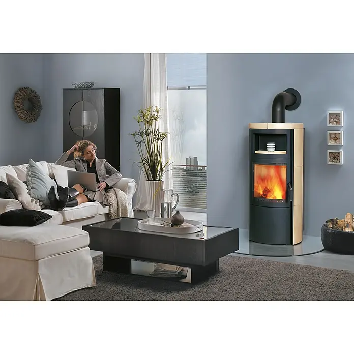 Hark Kaminofen VITO WW GT ECOPLUS - 14 kW, Raumheizvermögen: 116 m³, Material Verkleidung: Keramik, Creme-Weiss - Gemütliches Wohnzimmer mit einem Kaminofen mit Glasfenster, der eine angenehme Wärme ausstrahlt, und einer Frau, die auf der Couch sitzt.