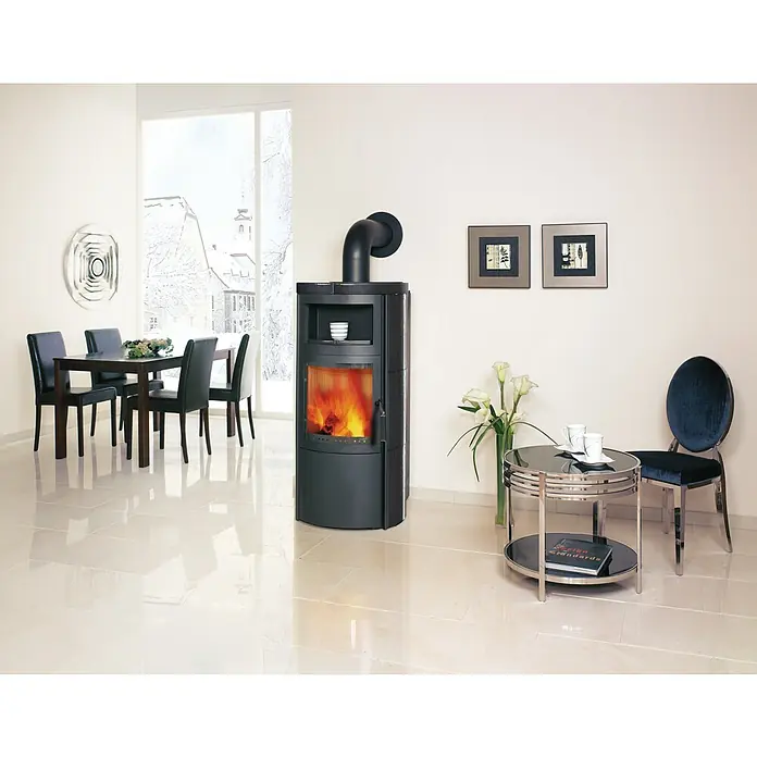 Hark Kaminofen VITO WW GT ECOPLUS - 14 kW, Raumheizvermögen: 116 m³, Material Verkleidung: Keramik, Meteor-Schwarz - Zimmer mit Kaminofen. Der Ofen ist schwarz und hat ein modernes Design. Im Hintergrund sieht man einen Esstisch und Stühle.