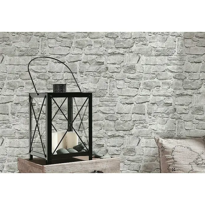 A.S. Création Il Decoro Vliestapete Bruchsteinmauer - Grau, Steinoptik, 10,05 x 0,53 m - Nahaufnahme einer schwarzen Laterne mit Kerzen und Steinen auf einem Holztisch vor einer grauen Steinwand.