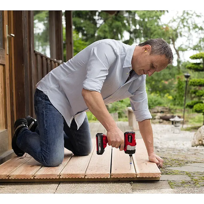Einhell Power X-Change 18V Akku-Schlagschrauber TE-CI 18/1 Li - 18 V, Ohne Akku - Ein Mann kniet und benutzt einen roten Einhell Akku-Schlagschrauber, um Holzplanken zu befestigen. Er trägt Ohrenschützer.