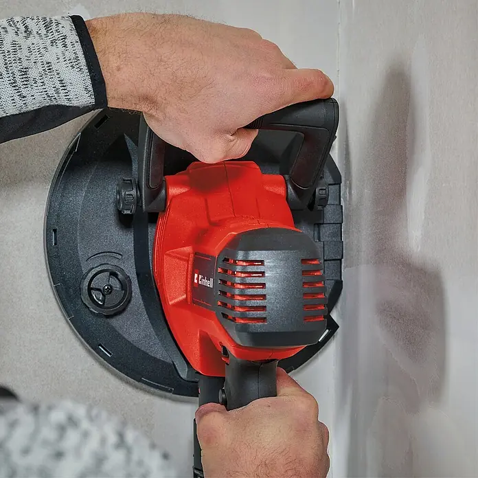 Einhell Trockenbauschleifer TC-DWS 225 - 1.220 W, Durchmesser Schleifteller: 215 mm, Leerlaufdrehzahl: 1.500 U/min - 2.300 U/min - Ein Mann benutzt eine Einhell Wandschleifmaschine zum Schleifen einer Wand. Das Gerät ist rot und schwarz und wird mit beiden Händen bedient.