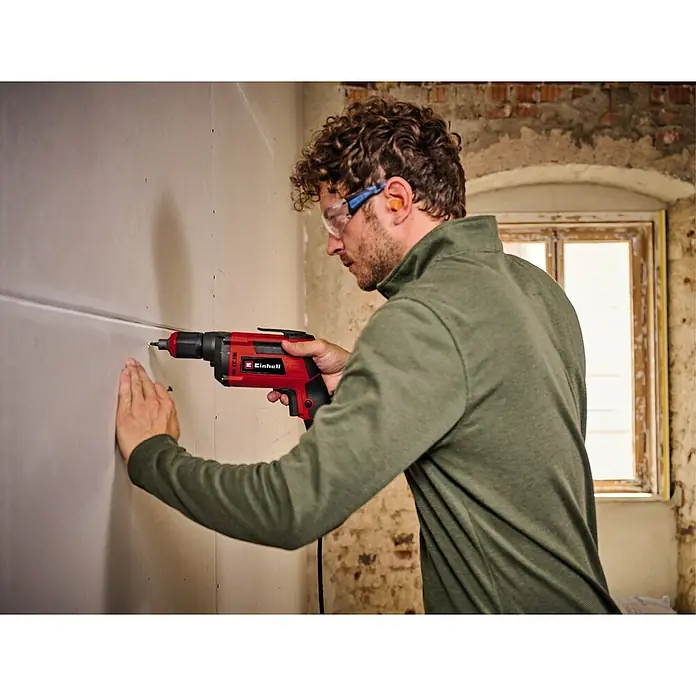 Einhell Classic Trockenbauschrauber TC-DY 710 E - 710 W - Ein Mann mit Schutzbrille benutzt einen roten Einhell Trockenbauschrauber an einer Wand.