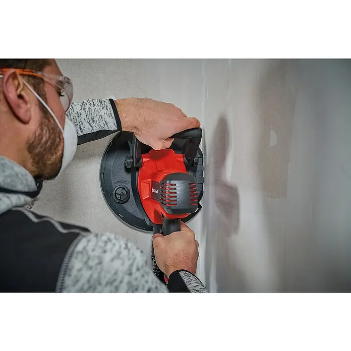 Einhell Trockenbauschleifer TC-DWS 225 - 1.220 W, Durchmesser Schleifteller: 215 mm, Leerlaufdrehzahl: 1.500 U/min - 2.300 U/min - Mann schleift Wand mit rotem Einhell Trockenbauschleifer, Schutzbrille und Maske für Sicherheit. Professionelles Werkzeug im Einsatz.