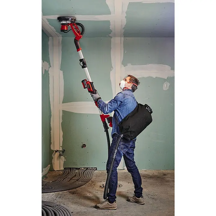 Einhell Professional PXC 18V Lijadora de pared y techo de batería TP-DW 18/225 Li - 18 V, Sin batería, Diámetro muela abrasiva: 225 mm - Un hombre usa una lijadora de paneles de yeso con aspiradora para lijar un techo. La herramienta es roja, plateada y negra.