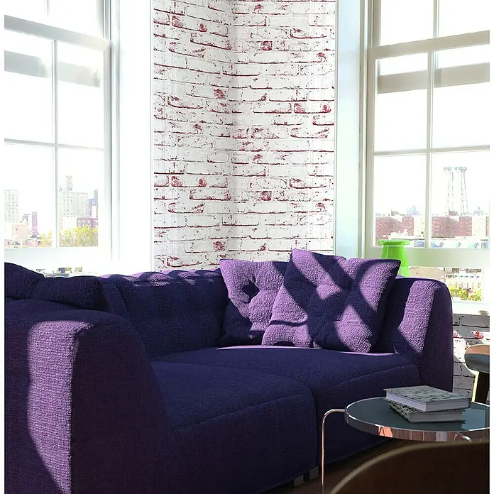 A.S. Création Casual Living Vliestapete New England Klinker - Weiß/Rot, Steinoptik, 10,05 x 0,53 m - Nahaufnahme eines modernen, lilafarbenen Ecksofas mit Kissen und einem Beistelltisch im Wohnzimmer.