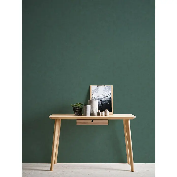 A.S. Création Casual Living Vliestapete - Grün, Uni, 10,05 x 0,53 m - Das Bild zeigt einen eleganten, hellen Holztisch mit einer Schublade, dekoriert mit Pflanzen, einem Bild und kleinen Keramikobjekten vor einer grünen Wand.