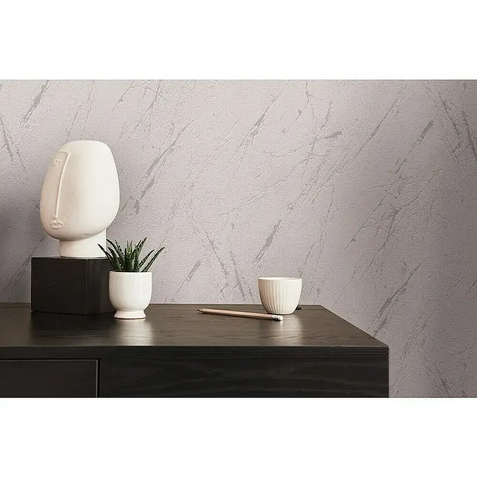 A.S. Création Casual Living Vliestapete Marmor - Silbergrau-Metallic, Steinoptik, 10,05 x 0,53 m - Nahaufnahme eines dekorierten Tisches mit einer weißen Skulptur, einer Pflanze, einer Tasse und einem Bleistift vor einer strukturierten Tapete.