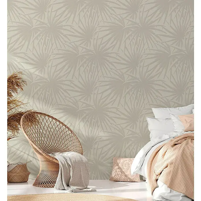 A.S. Création Casual Living Vliestapete Palmenblätter - Creme/Hellgrau, Floral, 10,05 x 0,53 m - Schlafzimmer-Interieur mit gemusterter Tapete, Rattan-Stuhl und Bett mit Kissen und Decke.