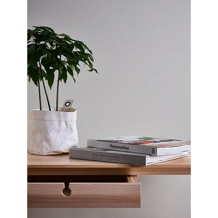 A.S. Création Casual Living Vliestapete - Strukturgrau, Uni, 10,05 x 0,53 m - Nahaufnahme eines Holzschreibtisches mit einer Zimmerpflanze in einer weißen Tasche und zwei Büchern, die eine gemütliche und minimalistische Ästhetik zeigen.