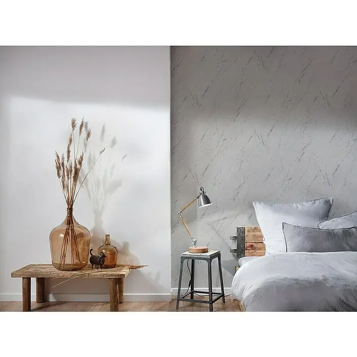 A.S. Création Casual Living Vliestapete Marmor - Silbergrau-Metallic, Steinoptik, 10,05 x 0,53 m - Eine große Vase mit getrockneten Blumen steht auf einem Holztisch neben einer Lampe auf einem Nachttisch in einem stilvollen Schlafzimmer.