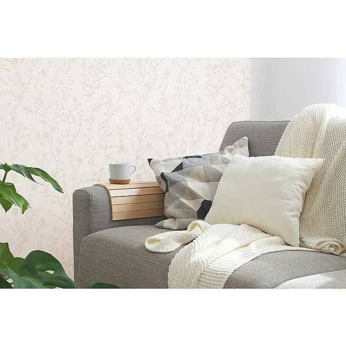 A.S. Création Casual Living Vliestapete - Hellrosa/Hellgrau, Floral, 10,05 x 0,53 m - Nahaufnahme eines Sofas mit einer Holzarmlehne, einer Tasse, Kissen und einer Decke. Gemütliches Interieur mit dekorativer Tapete im Hintergrund.