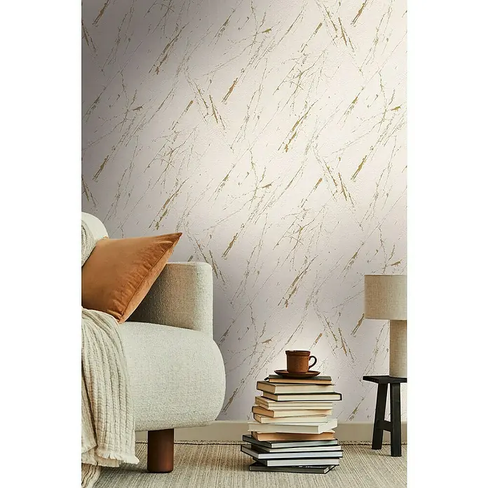 A.S. Création Casual Living Vliestapete Marmor - Beige/Gold/Metallic, Steinoptik, 10,05 x 0,53 m - Nahaufnahme eines Sofas mit Kissen, einer Bücherstapel und einer Lampe vor einer Wand mit abstraktem Marmormuster.