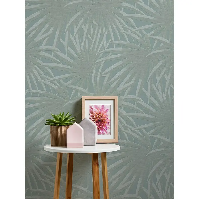 A.S. Création Casual Living Vliestapete Palmenblätter - Blau/Mint, Floral, 10,05 x 0,53 m - Nahaufnahme eines kleinen runden Beistelltisches mit Topfpflanze, Dekohäusern und einem gerahmten Foto auf einem stilvollen Hintergrund.